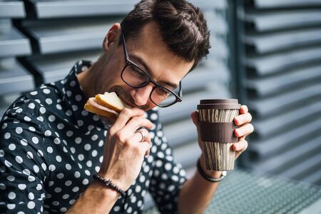 Young businessman sitting outdoors, drinking coffee and eating sandwich.の写真素材