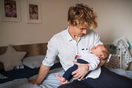A young father with a newborn baby son sitting on bed at home.