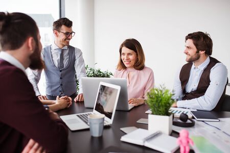 A group of young business people with laptop sitting in an office, talking.