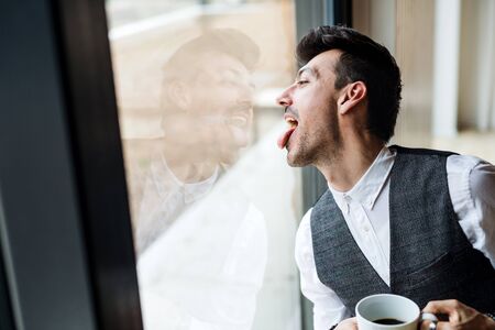 Young man with coffee standing by the window, sticking out tongue.の写真素材