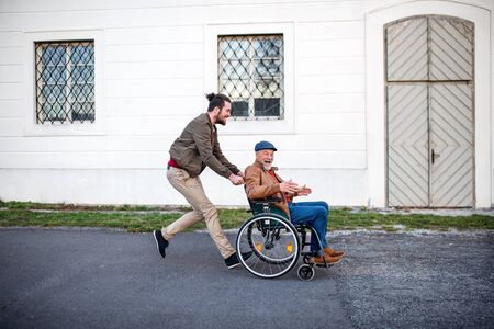 Young man and his senior father in wheelchair on a walk in town, having fun.の写真素材