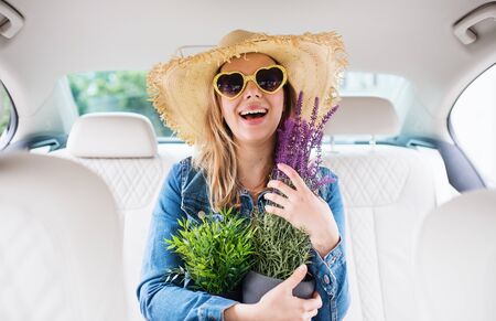 Young woman with hat and sunglasses sitting in car, holding plants.