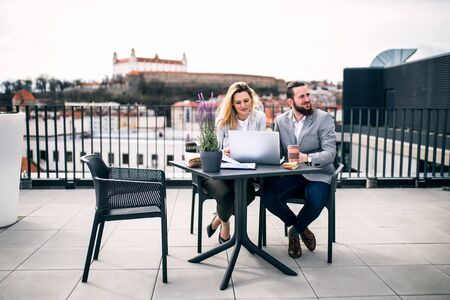 Two young business people with laptop sitting on a terrace outside office, working.の写真素材
