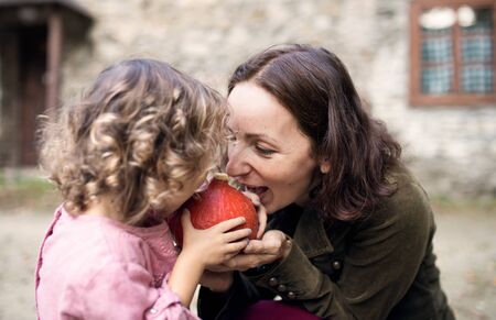 A small girl with mother standing in front of old stone house, holding pumpkin.の写真素材