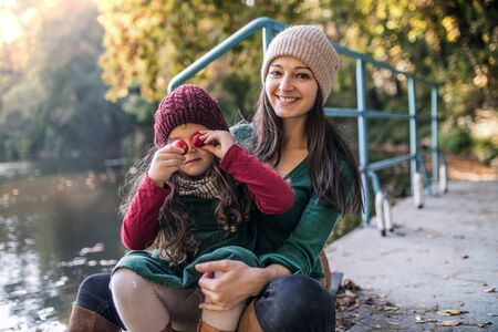 A young mother with a toddler daughter sitting in forest in autumn nature.の写真素材