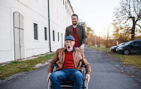Young man and his senior father in wheelchair on a walk in town, having fun.の写真素材