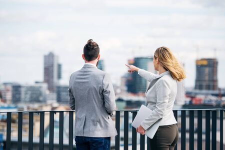 Two young business people with laptop standing on a terrace outside office, talking.の写真素材
