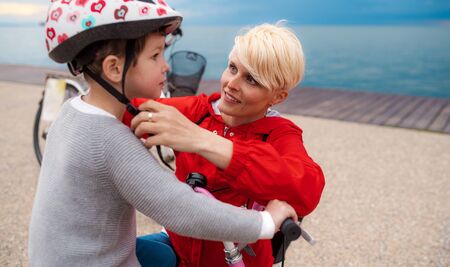 Mother and small daughter with bicycle outdoors on beach, putting on helmet.の写真素材
