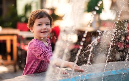 Small girl standing by water fountain outdoors in town.の写真素材