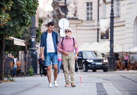 Young man and blind senior with white cane walking on pavement in city.の写真素材