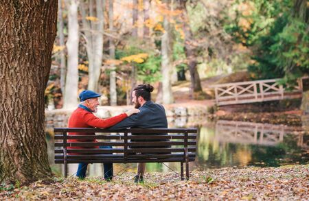 Rear view of senior father and his son sitting on bench by lake in nature, talking.の写真素材