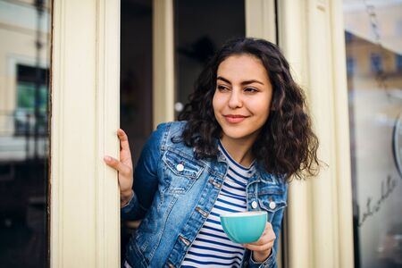 A beautiful young woman with tablet and coffee standing at the front door in cafe.