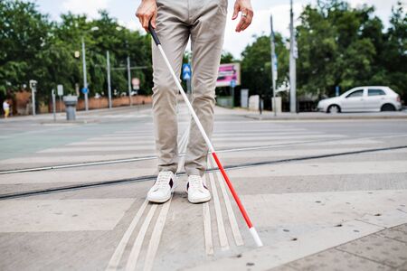 Midsection of young blind man with white cane walking across the street in city.の写真素材