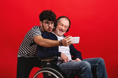 Young man and senior father in wheelchair in a studio on red background, taking selfie.の写真素材
