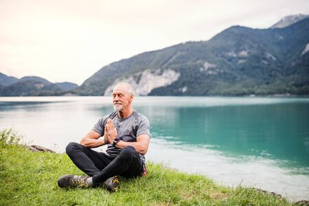 A senior man pensioner sitting by lake in nature, doing yoga exercise.の写真素材