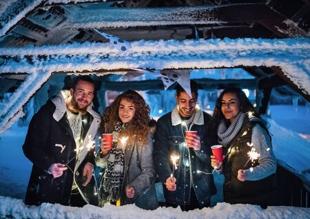 Group of young friends outdoors in snow in winter at night, holding sparklers.