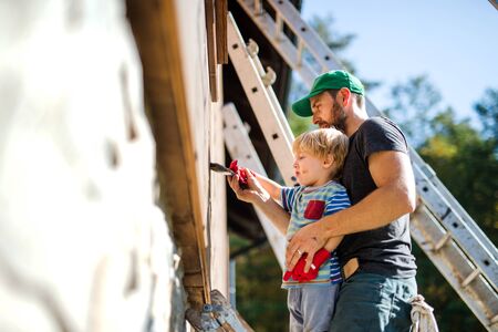 A father and toddler boy outdoors in summer, painting wooden house.の写真素材