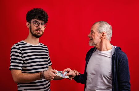 Senior father giving money to his son in a studio on red background.