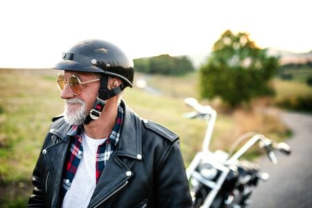 A front, view portrait of cheerful senior man traveller with motorbike in countryside, walking.の写真素材