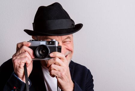 Portrait of a senior man with hat and camera in a studio, taking photograph.