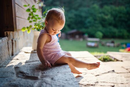 A cute toddler girl sitting outdoors in front of house in summer.の写真素材