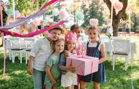 Portrait of small girl with friends and present outdoors in garden in summer.