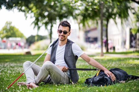 Young blind man with white cane and guide dog sitting in park in city.