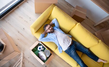 Top view of mature man with boxes moving in new house, relaxing on sofa.