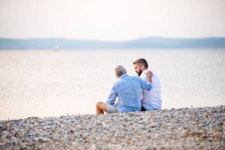 Rear view of senior father and mature son sitting by the lake, talking.の写真素材
