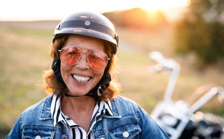 A cheerful senior woman traveller with motorbike in countryside at sunset.の写真素材