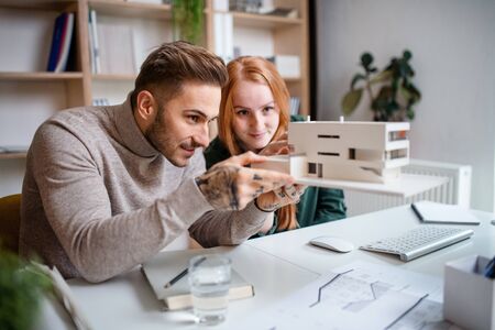 Architects with model of house sitting at the desk indoors in office, working.