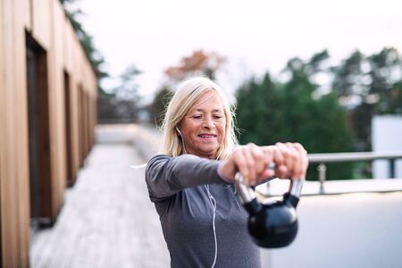 A senior woman with earphones and kettlebell outdoors doing exercise.の写真素材
