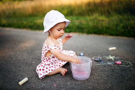 Cute toddler girl outdoors in countryside, chalk drawing on road.