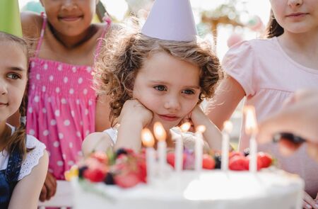 Children with cake standing around table on birthday party in garden in summer.