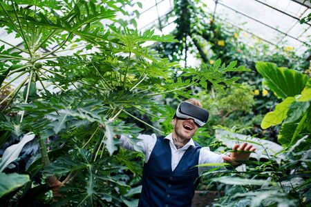 Front view of young man standing in botanical garden, using VR glasses.
