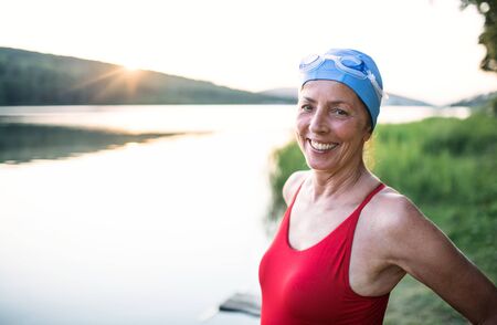 Senior woman in swimsuit standing by lake outdoors before swimming.の写真素材