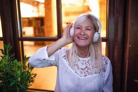 Senior woman with headphones standing outdoors on terrace, listening to music.