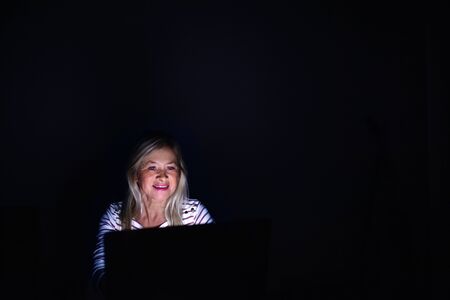 Senior woman with laptop indoors at home working late at night.