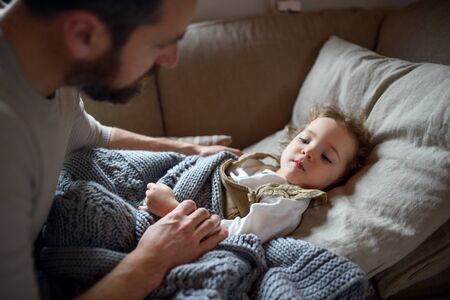 Father checking on small sick daughter lying in bed indoors at home.