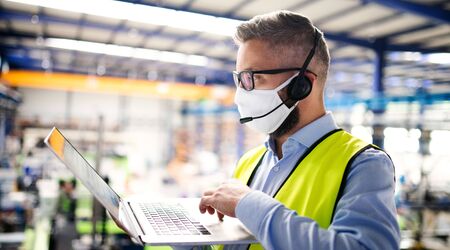 Technician or engineer with protective mask and laptop working in industrial factory.