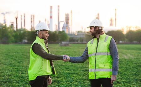 Two young engineers standing outdoors by oil refinery, shaking hands.の写真素材