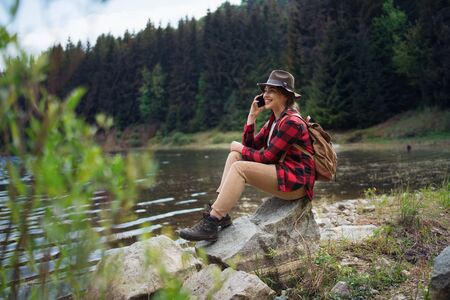 Young woman sitting by lake on a walk outdoors in summer nature, using telephone.