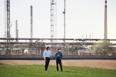 Two young engineers standing outdoors by oil refinery, discussing issues.の写真素材