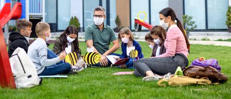 Group of cheerful children with face masks learning outdoors at school after lockdown.