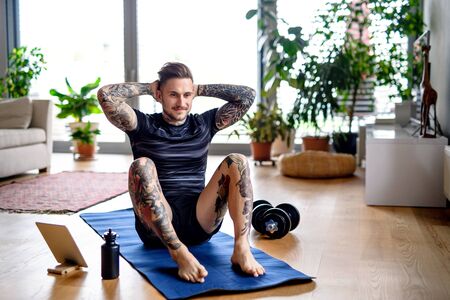 Front view portrait of man with tablet doing workout exercise indoors at home.