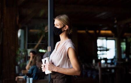 Side view of waitress with face mask standing at the door in restaurant.の写真素材