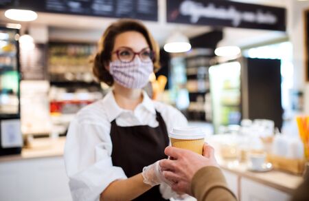 Young woman with face mask working indoors in coffee shop, serving customer.の写真素材