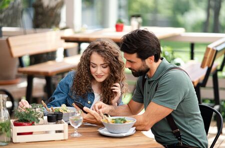 Happy couple sitting outdoors on terrace restaurant, using smartphone.の写真素材