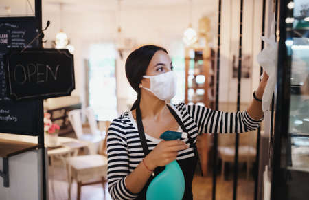 Young woman with face mask working indoors in cafe, disinfecting surfaces.の写真素材