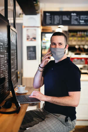 Young man with face mask and laptop indoors in cafe, using smartphone.の写真素材
